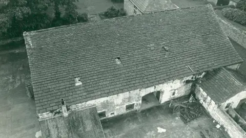 National Trust An archive black and white aerial image shows the barn in the 1980s. It has a large sloping rectangular roof with a few patches of missing tiles and two attached smaller extensions to the front left and front right. There is an open archway in the middle.