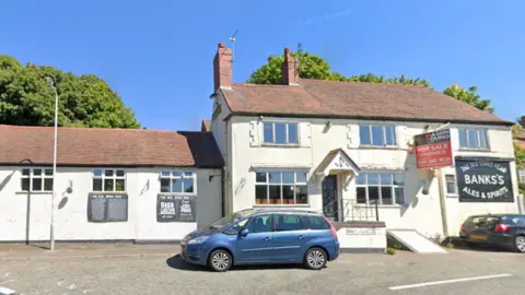 The pub is white and has an adjoining building. There is a for sale sign up with two cars parked outside the front.