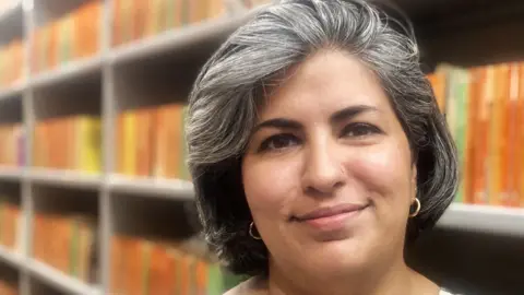 Mousumi Bakshi/BBC Zainab Juma stands in front of shelves of mostly orange books. She has grey, swept-back hair and gold earrings and is smiling.