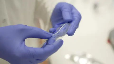 The device known as SilkAxons is being held in the hands of Alex Woods who is wearing blue gloves. The device is made of multiple spider silk fibres.