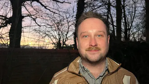 A smiling man with a short beard smiling at the camera at dusk. There are trees in the background