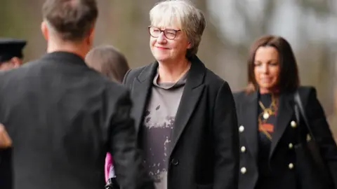 PA Media Susan Stewart has short, grey hair and glasses. She is wearing a grey dress with a burgundy pattern on it and a black jacket over the top. She is smiling at a man who has his back to the camera. There are other mourners behind her.