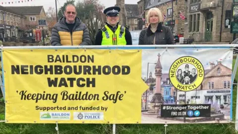 A police officer and two members of the public stand behind a sign on a grassed roundabout, which reads 'Baildon Neighbourhood Watch: Keeping Baildon Safe'.