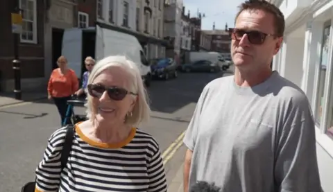 A woman with shoulder length hair and a striped top standing next to a man with sunglasses and a grey t-shirt. There are other people and a van in the background.