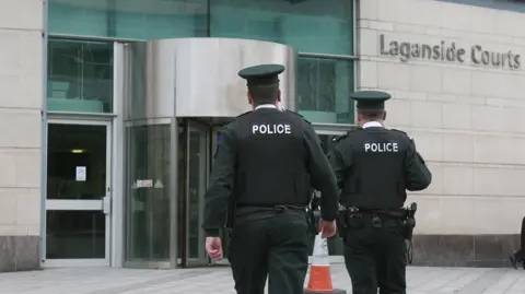 PA Media Two male police officers walk towards a cream brick building which has a green entrance and silver revolving doors. On the cream wall to the right of the entrance there is a sign which reads ' Laganside Courts'. A man in a black suit leans on a black bin smoking a cigarette. 