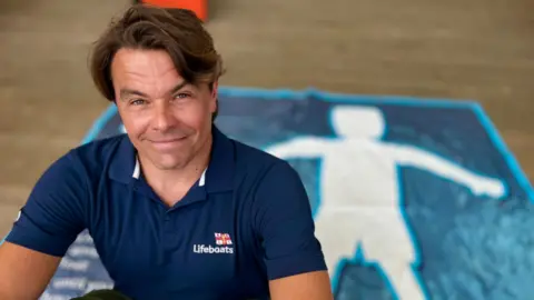 Martin Giles/BBC A head and shoulders shot of Nick Ayers, who has mid-brown hair and is wearing a navy blue RNLI polo shirt and is starting to smile. He is sitting down with his arms around his knees and behind him is a blurred-out blue mat with the white outline of a human figure on its back with its arms and legs outstretched.