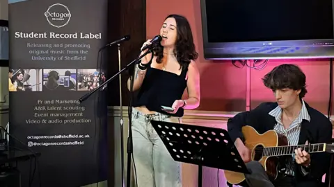 A singer and guitarist perform. A banner that reads 'Student Record Label' is positioned next to the singer.