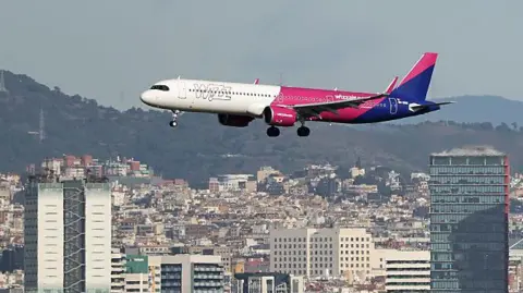 A white, pink and blue airplane with "Wizz" written on the side flies low over a city, with wooded hills in the background.