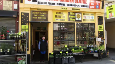 Garston Garden Centre/Facebook Garston Garden Centre shop front is painted yellow with flowers and plants outside. Lill is stood in the doorway smiling