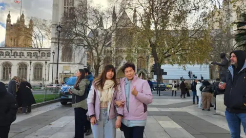 Two women standing in Parliament Square. In the background, there is Westminster Abbey and the Victoria Tower of the Houses of Parliament.  