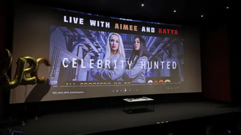 Getty Images A huge projector says: Live with Aimee and Katya. Celebrity Hunted. There is a stage just below it.