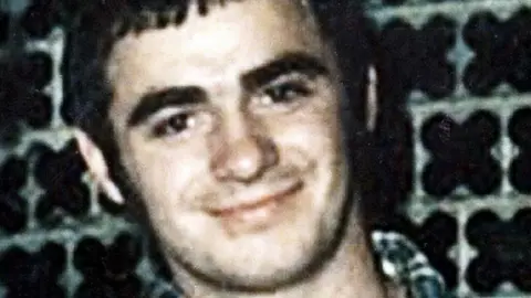 Pacemaker A colour picture of Danny McIlhone. He is standing against patterned wallpaper and has dark eyebrows and short dark hair. He is wearing a navy patterned shirt and has a smiling face looking directly to camera.