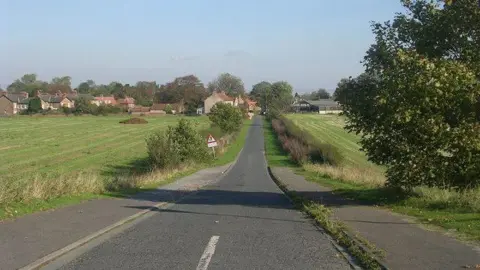 A country lane with fields either side and housing in the distance.