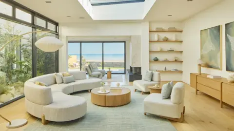 Omaze A lounge with a large window with sea views. There is a light-coloured rounded sofa and matching chairs, along with a wooden coffee table and sideboard.