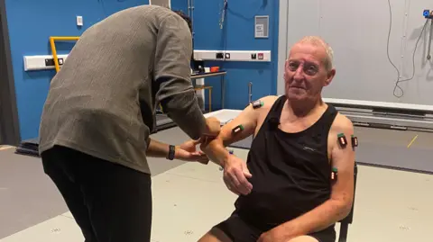 BBC Bernie Kielty is sat down in a large room. He is wearing a black vest and has lots of different technology and equipment attached to his arms. There is a man in a grey top looking at his right arm.