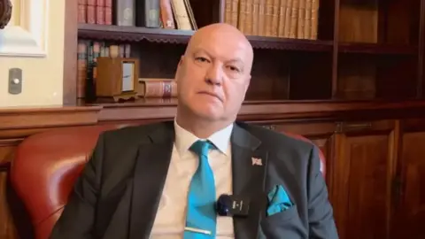 BBC Ian Cooper sits in a maroon leather chair, with wooden panelling and a recessed bookcase behind him. He is wearing a white shirt, dark grey suit jacket, with a union flag pin badge and a clip microphone on the lapel, and a teal blue tie. He is looking into the camera with a serious expression.