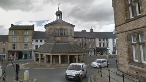 Google Market Cross is an octagonal stone structure at the centre of a roundabout. Columns line the very bottom and the top and there is a weather vane at the very top. The shops lining the surrounding streets are terraced.