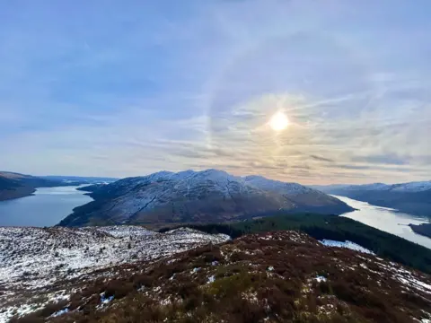 Charlie McGinn Looking down at snow-capped mountains with a loch on either side. The sun is shining and there is a snow bow around it