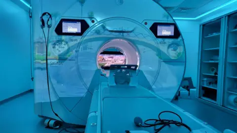 The scanner has two screens on the outside and a screen can also be seen inside at the back. A table has a support for the patient's head. There is relaxed blue lighting in the room.