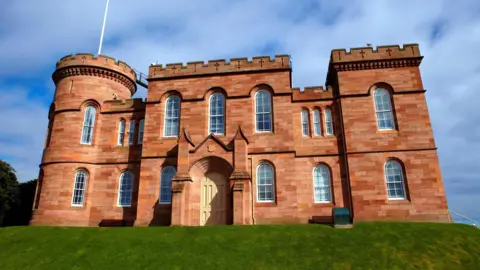 A view looking up at Inverness Castle. It is a sandstone building with turrets and battlements. There is an area of cut grass in front of the castle.
