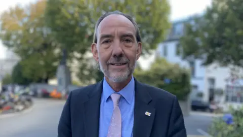 Man wearing a Navy suit, blue shirt and a light beige pinkish tie. The man has a small brooch pin of the Guernsey flag.