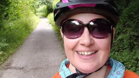 A smiling Pippa Marshall is wearing a bike helmet and orange T-shirt with the MS Society logo, and dark glasses.