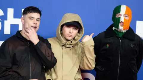 Getty Images Three young men stand next to each other on a red carpet. One has his hand on his face. The middle man is pointing towards the last man, who is wearing a green , white and orange balaclava over his face. 