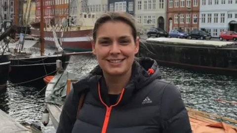 Family handout Tarryn Baird smiles at the camera standing in front of Copenhagen docks with sail boats and colourful buildings behind her