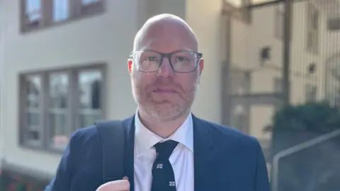 BBC Deputy Rob Curgenven - A man with a bald head, a ginger and grey beard, wearing a blue suit, and a white shirt, with a blue and Guernsey flag logo tie. 