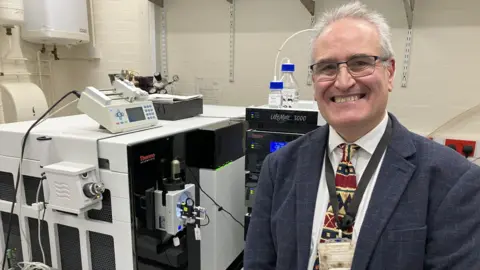 BBC Professor Mur is pictured in a laboratory with a number of large and small machines behind him. He is wearing a grey jacket, white shirt and patterned tie and is smiling at the camera. He has short grey hair.
