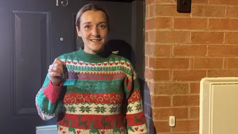 A woman in a Christmas jumper smiling outside a dark grey door. In her right hand she is holding a key, to her right is brickwork and a doorbell illustrating she is standing in front of a house.