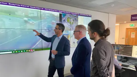 Three men looking at a projection of CCTV footage of a road. They are all standing side-on and wearing business wear. The CCTV footage illustrates a car pulling out of a junction. They are standing in an office.  