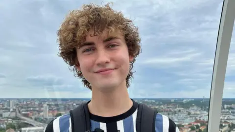 Ethan Brown looking directly at the camera. He has curly brown hair and is wearing a t-shirt with blue, black and white vertical stripes, and has a rucksack on his back. In the background is a view over a city.