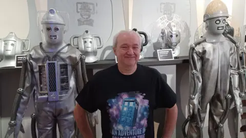 Paul Hayes/BBC A grey-haired man in his late 50s, smiling into the camera with his hands on his hips. He is wearing a black T-shirt with a picture of a blue police box on it, and the caption ‘An Adventure in Space and Time’. Behind him, on display as exhibits, are two Cybermen costumes, robotic-looking alien villains from the BBC science-fiction series Doctor Who.