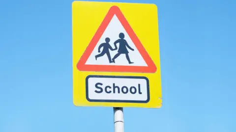 Getty Images A yellow road sign which has a red triangle on it and a picture of two people holding hands and crossing the road. Underneath the triangle there is a rectangle with the word school written in it in black letters.