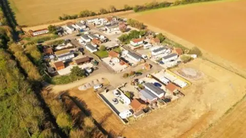 An aerial view of the Pines Residential site in Frome, Somerset. The site has 25 pitches with caravans and trailers. It is surrounded by golden brown fields and green trees.