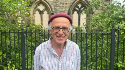 Jessica Bayley/BBC A man in his late 60s wearing a white and pastel checked shirt and a red beret and glasses smiling whilst stood in front of a steel fence in front two arches covered by greenery.