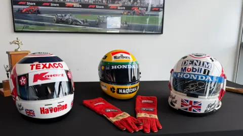 Robert Constantin/BBC Three helmets and a pair of red racing gloves displayed on a table. One of the helmets has been signed.