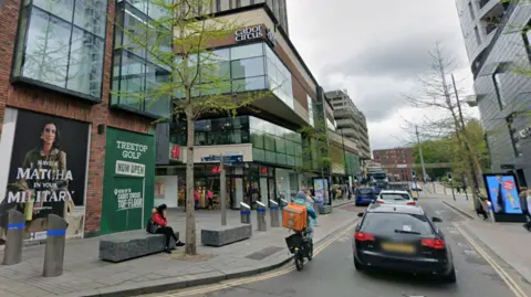 A street view of a main street running through Bristol, with a shopping centre on one side. There is traffic and a bike passing, and high-rise buildings on either side. 