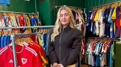 An 18-year-old girl with long blonde hair is looking directly at the camera as she's stood in her store surrounded by hundreds of old football shirts 