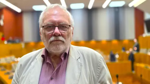 Paul Moseley/BBC Steve Morphew is wearing a light, cream linen jacket over a red and white pin-striped shirt. Behind him, there is a council chamber.