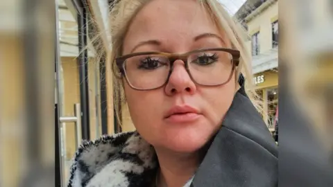 A head and shoulders shot of Gemma Whitehead. She has blonde hair tied back and is wearing brown glasses. She is wearing a grey coat and a black and white scarf. 