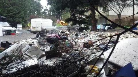 Rubbish, including paper, clothes and white goods, is piled up around knee-high on a tarmac courtyard next to a white building. The area is bordered by tall trees. There are two white vans parked on the courtyard too. 