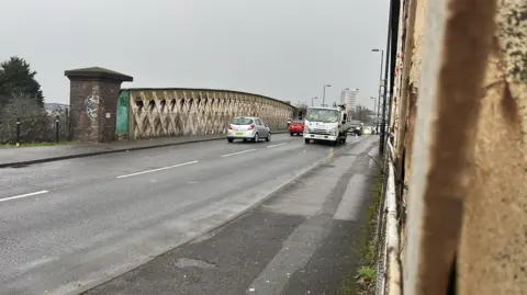 A road along an old looking rail bridge with cars travelling along it.