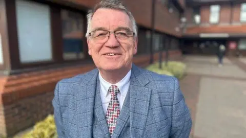 Martin Gannon smiling at the camera. He has grey, short hair and is wearing glasses, a grey checked suit and colourful tie.