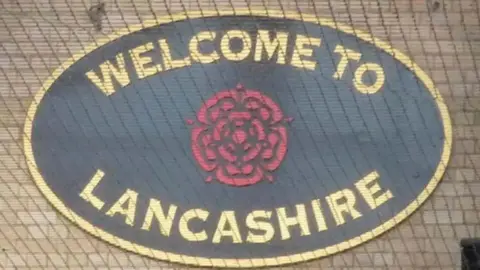 Oval sign saying "welcome to Lancashire" with a red rose in the middle. the writing is gold on a black background