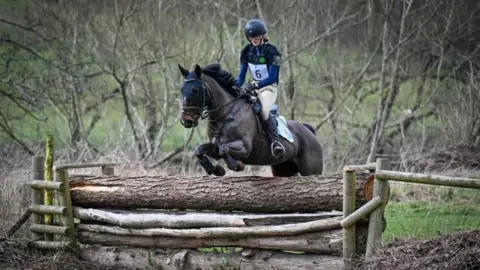 Courtfield Estate A black horse ridden by a girl in beige jodhpurs, a blue top and a black hat leaps over a log pile.