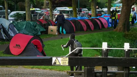 PA Media Early morning work to remove tents pitched by asylum seekers along the Grand Canal, Dublin. There are lots of tents and people in jackets and a garda can be seen in the background.