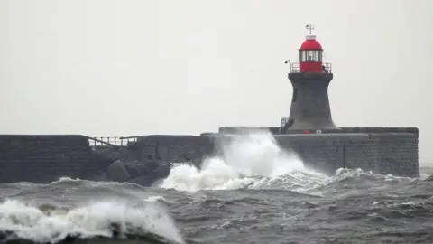 Damage is visible to the pier structure which has large rocks in front of it. There is a lighthouse, large waves and three birds in flight.