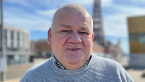 A close-up of Ian White, whose hair is shaved short. He is wearing a shirt and a blue crew-neck jumper. The blurred background features the promenade in Blackpool including the Blackpool Tower